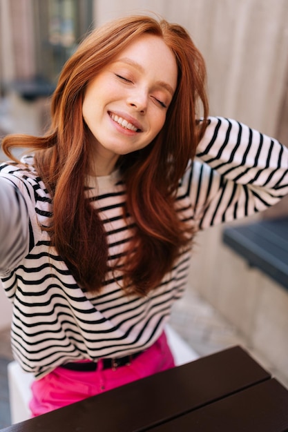 Vertical POV portrait of smiling woman with closed eyes taking self portrait with smartphone sitting at table in outdoor cafe Point of view of redhead female doing selfie or having online video call