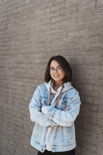 Vertical protrait of an attractive young girl with short hair, wearing glasses and street style clothes, leaning on a brick wall.