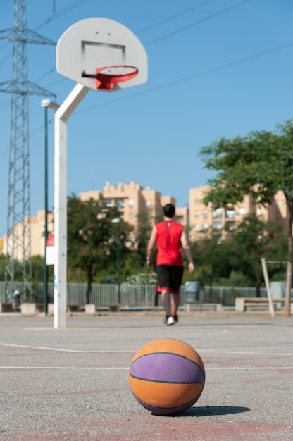 Вертикальный снимок с выборочным фокусом баскетбольного мяча на площадке с игроком-мужчиной, идущим рядом с рингом