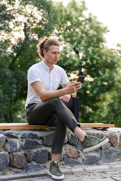 Vertical Serious curly business man with briefcase using smartphone while sitting on bench in park