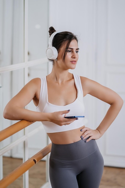 Vertical shoot of brunette hispanic girl in sportswear at break of workout at gym using headphones phone enjoys music Fit caucasian model with perfect body shape at exercise Fitness weight loss