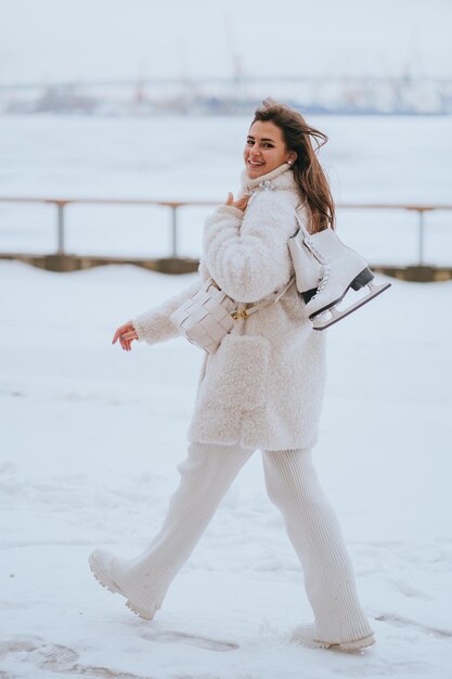 Photo vertical shoot of caucasian brunette girl in white fur coat white pants carrying skates