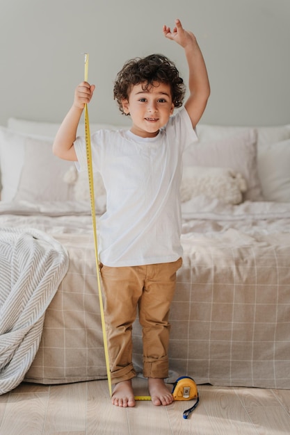 Vertical shoot of curly handsome Spanish boy measuring himself by measuring roulette standing at bedroom Kids growing concept Healthy people and childhood Pretty preschooler checking his height