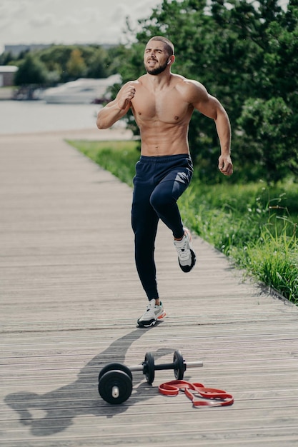 Vertical shot of active European musular man jumps and does sport outdoor has cardio training listens favorite music in earphones breathes fresh air uses sport equipment has naked torso