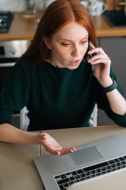 Vertical shot of arguing angry young woman talking on mobile phone and using laptop at table in kitchen with modern interior Annoyed female freelancer talking on cellphone having problems with job