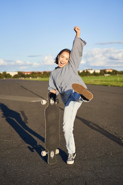Vertical shot of asian girl feeling excited skating on longboard jumping and posing with skateboard