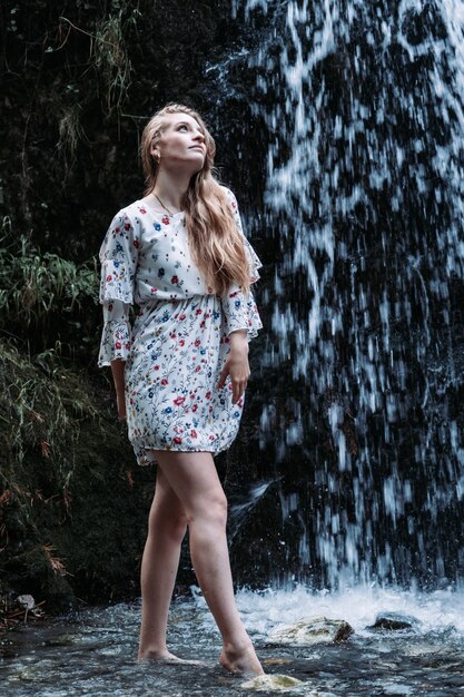 Vertical shot of an attractive young woman in a light summer dress posing with a waterfall behind