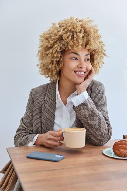 Vertical shot of beautiful elegant young woman with curly hair\
dressed in formal clothes holds cup of coffee eats croissant poses\
at wooden table with smartphone isolated over white background