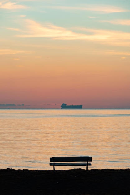 Вертикальный снимок скамейки на берегу моря на фоне закатного неба