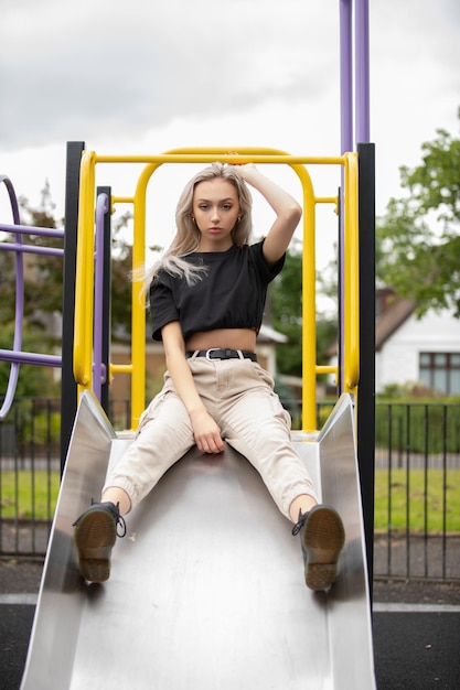 Vertical shot of a blonde Caucasian girl sitting on a slide in the background of the buildings