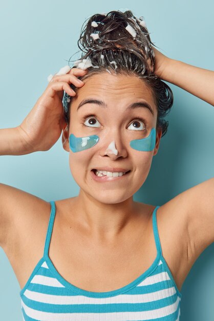 Vertical shot of brunette asian woman washes head with shampoo\
applies beauty patches under eyes to reduce wrinkles takes shower\
wears striped t shirt isolated over blue background hygiene\
concept