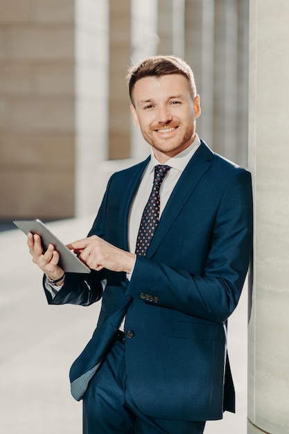 Photo vertical shot of business person uses digital tablet for online communication and work uses free internet at office has positive expression dressed in formal black suit people and occupation