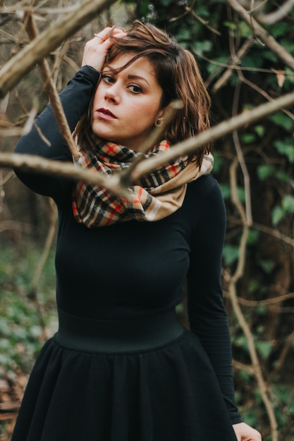 Vertical shot of a Caucasian female dressed in a black dress and a scarf in the forest in autumn