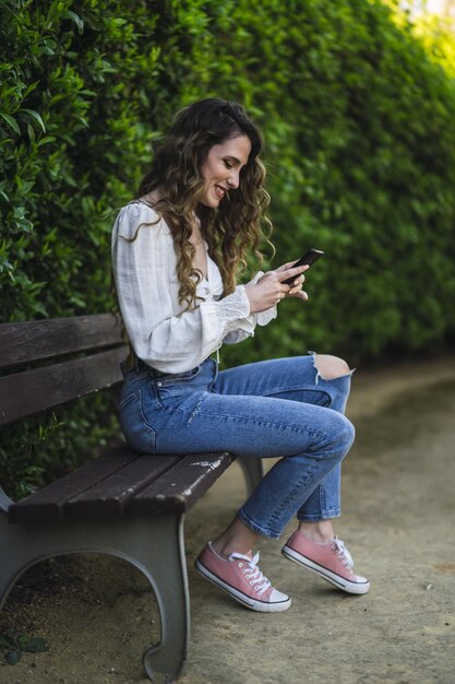 彼女の携帯電話のベンチテキストメッセージに座っている白いトップのスペインからの白人女性の垂直ショット