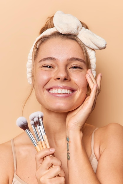 Vertical shot of cheerful European woman touches face gently smiles toothily holds cosmetic brushes has natural beauty smooth healthy clean skin wears headband isolated over beige background