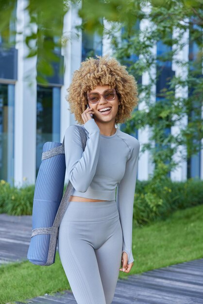 Vertical shot of cheerful fit womn makes phone call via\
smartphone wears sunglasses and tracksuit carries rolled fitness\
mat going to practice yoga outdoors has happy expression stands in\
city park