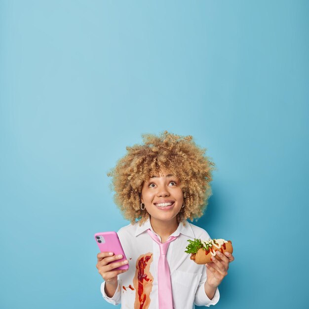 Vertical shot of cheerful young woman with curly hair focused\
above gladfully holds mobile phone and delicious hot dog eats cheat\
meal has addiction to fast food isolated over blue background