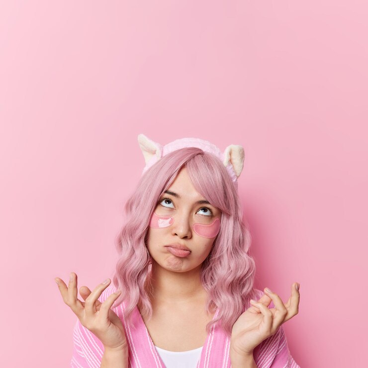 Vertical shot of clueless hesitant woman spreads palms looks sadly above cannot make decision applies hydrogel patches under eyes to reduce wrinkles isolated over pink background copy space.