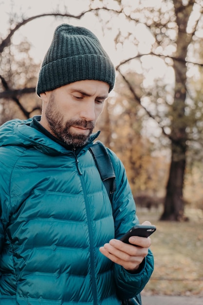 Vertical shot of concentrated hipster guy with thick beard wears anorak and hat holds cellular uses special application for finding route message with friend breathes fresh air Lifestyle concept