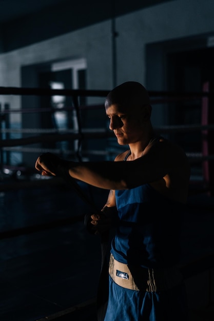 Vertical shot of confident boxer wrapping defense boxing tape\
around arms before fight in sport club with dark interior on\
background of ring. serious fighter getting ready for fight in\
fitness center