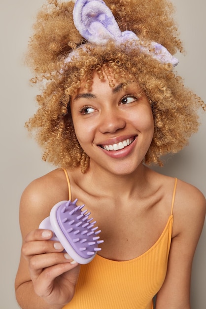 Vertical shot of curly haired cheerful woman wears headband and casual t shirt holds body massager has healthy clean skin smiles toothily isolated over grey background Daily routines concept