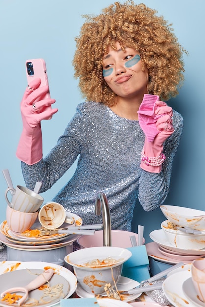 Vertical shot of curly haired housewife washes dirty dishes\
after party poses at smartphone front camera wears rubber gloves\
applies hydrogel patches under eyes wears festive dress blue\
background