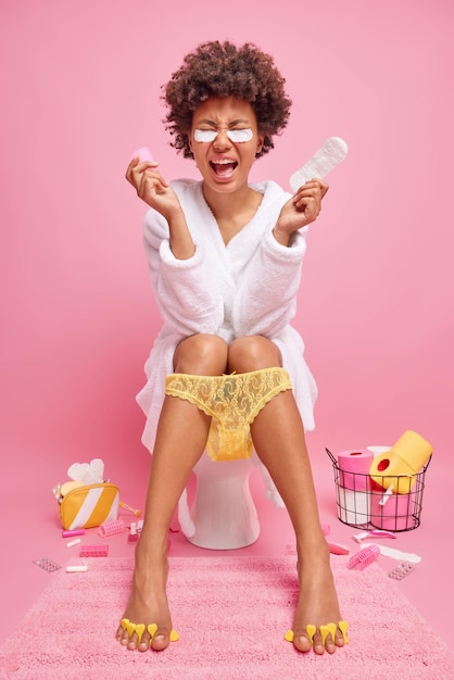 Photo vertical shot of curly haired woman holds menstrual cup and sanitary napking for menstruation wears bathrobe and lace panties pulled down on legs poses on toilet bowl isolated over pink wall