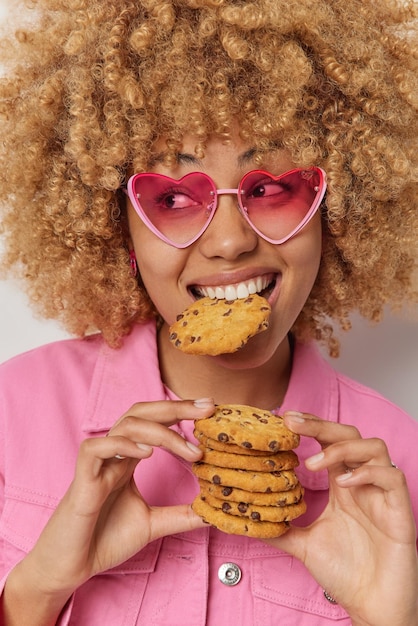 Photo vertical shot of curly haired young woman eats delicious cookies wears heart shaped sunglasses and pink jacket has unhealthy nutrition looks away with cheerful facial expression lunch time