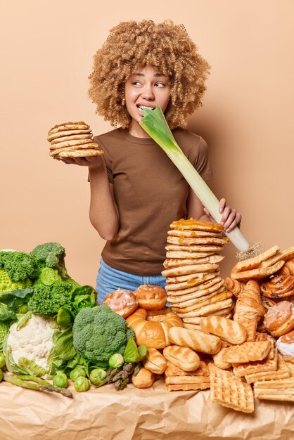 Vertical shot of curly woman bites leek tries to keep healthy\
diet feels temptation to eat unhealthy sweet food dressed in casual\
t shirt isolated over beige background binge eating over organic\
one