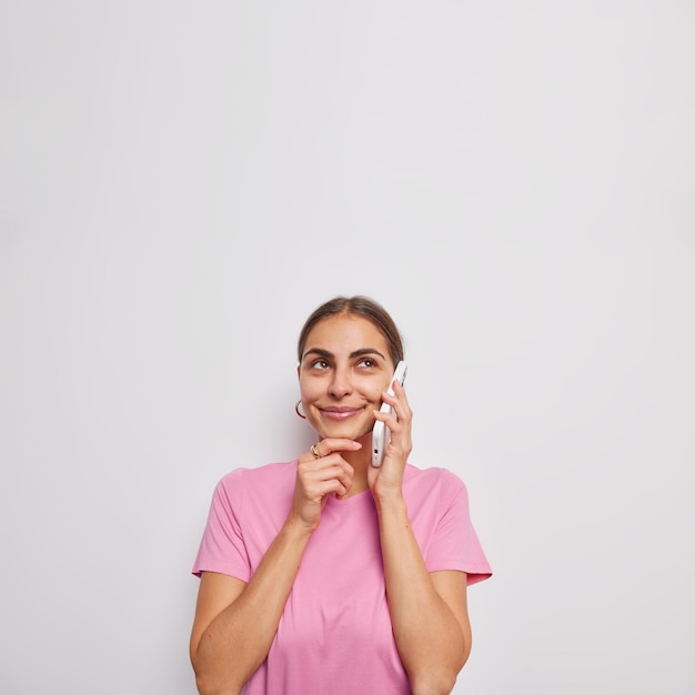 Il colpo verticale della donna premurosa sognante tiene il mento concentrato sopra ha la conversazione telefonica tiene il telefono cellulare vicino all'orecchio indossa una maglietta rosa casual isolata su sfondo bianco spazio vuoto