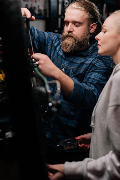 Вертикальный снимок сфокусированного бородатого мастера по ремонту велосипедов, общающегося с клиенткой, показывающей проблему с велосипедом, обнаруженную во время диагностики в ремонтной мастерской