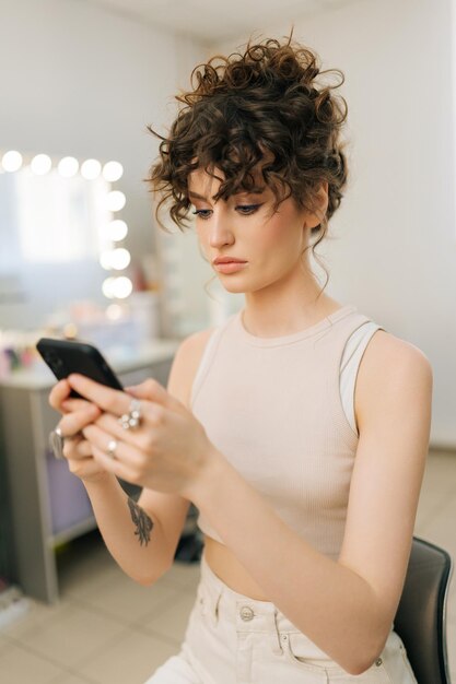 Vertical shot of focused pretty young woman with perfect makeup and hairstyle typing online message using smartphone looking to screen having cosmetic beauty care hair face procedure in visage studio