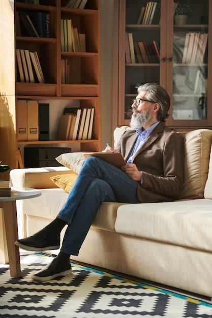 Vertical shot of handsome bearded man in stylish outfit with eyeglasses sitting on sofa in modern office room holding clipboard looking away