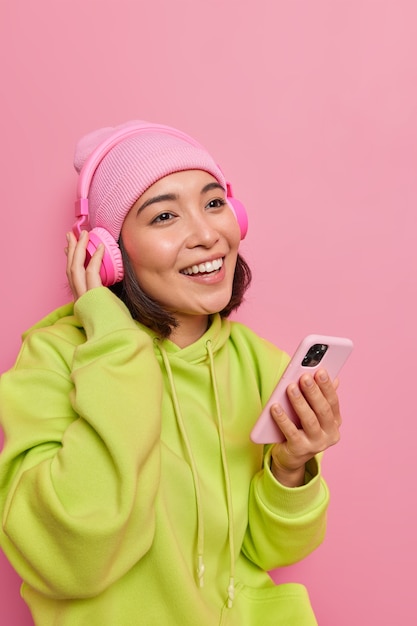 Vertical shot of happy Asian teenage girl holds mobile phone in hand enjoys listening music from playlist has pleasant smiles on face dressed casually