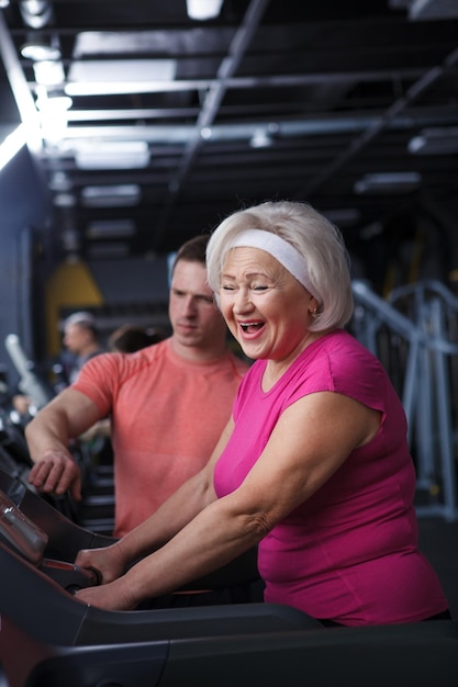 Ripresa verticale di una donna anziana felice che ride mentre si allena con il personal trainer in palestra camminando su un tapis roulant