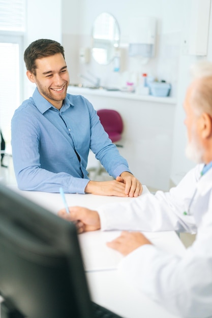 クリニックオフィスでの健康診断の訪問中にヘルスケアのアドバイスを与えるシニア男性医師を聞いている幸せな若い男性患者の垂直ショット。病院で笑顔の若い男にインタビューする主治医。