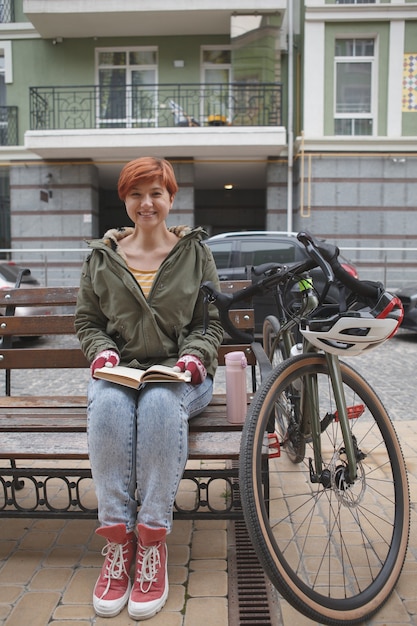 Vertical shot of a happy young woman laughing, reading a book in city center, resting after cycling