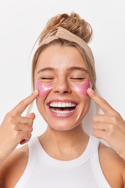 Vertical shot of happy young woman points at pink hydrogel patches under eyes smiles broadly shows perfect white teeth wears headband undergoes beauty treatments remves wrinkles or puffiness