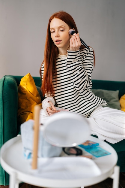 Vertical shot of injured young woman with broken right arm wrapped in white gypsum bandage applying cosmetic on face with professional makeup brush