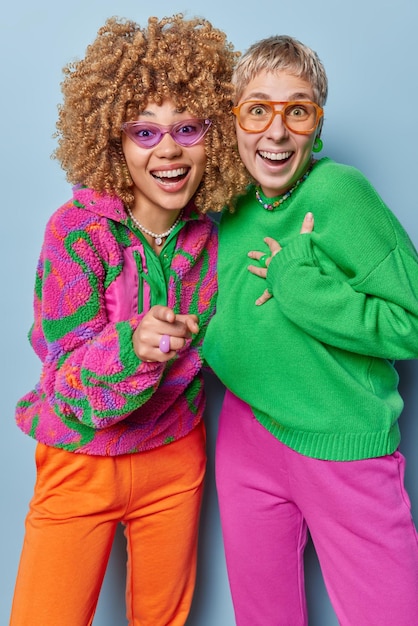 Vertical shot of joyful women react to something funny point\
directly at camera stand next to each other dressed in colorful\
jumpers and trousers isolated over blue background happy\
emotions