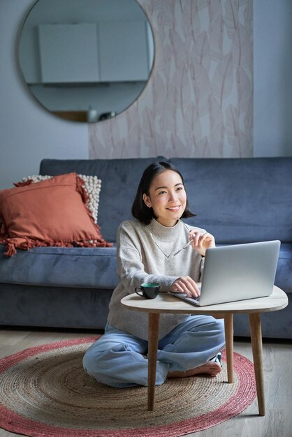 Vertical shot of korean working woman sitting on floor at home with laptop studying using computer o