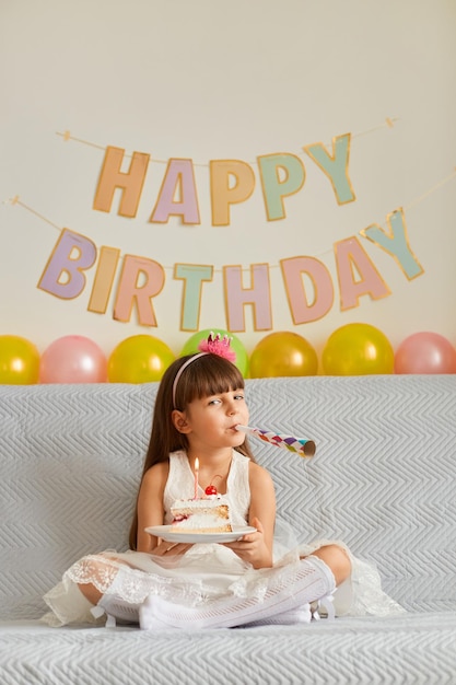 Vertical shot of little female child wearing white dress sitting on sofa, looking at camera with party horn, celebrating birthday, posing with balloons and festive inscription on background.