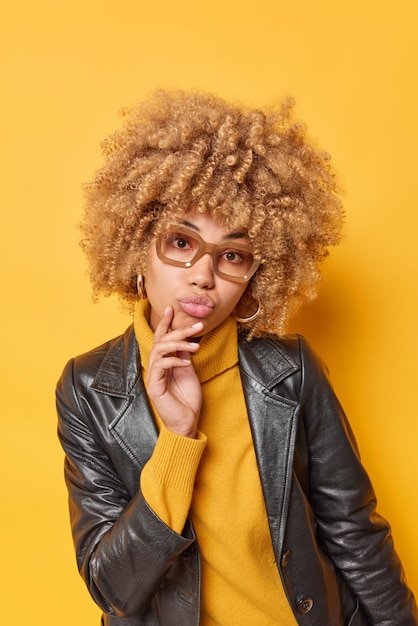 Vertical shot of lovely curly haired young woman keeps lips folded has romantic face expression wears spectacles stylish leather jacket isolated over yellow background. Human expressions concept