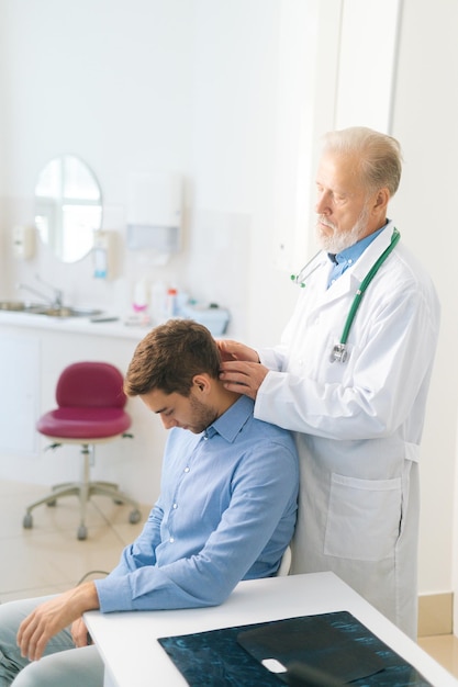 Vertical shot of mature adult male manual therapist turning and bending head of male patient to check his injured neck. Young man on preventive examination of doctor chiropractor on neck diagnostic.