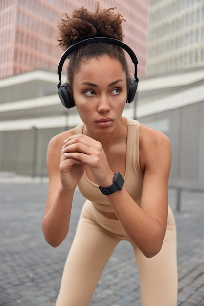 Vertical shot of motivated sporty woman keeps hands together dressed in sportswear makes squatting exercises concentrated away with thoughtful expression listens music via wireless headphones