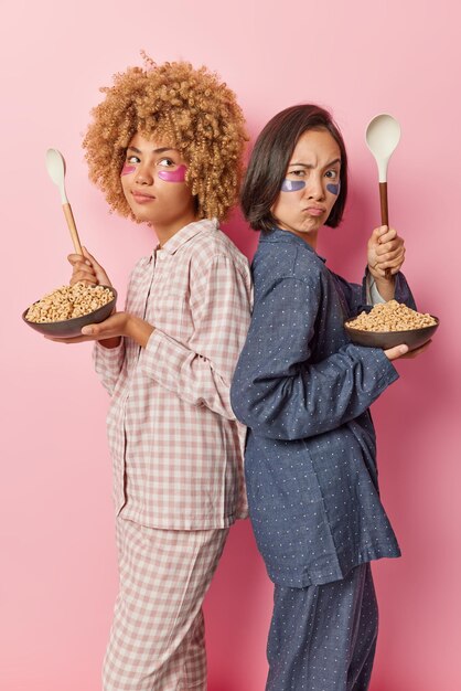 Vertical shot of multiethnic women stand backs hold bowls of\
cereals and big spoons going to have healthy breakfast dressed in\
nightwear apply hydrogel patches under eyes to moisturize skin