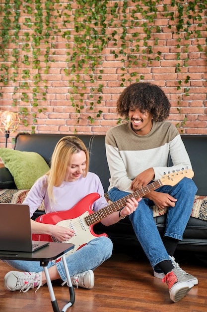 Vertical shot of multiracial couple sitting on sofa at home learning to play electric guitar