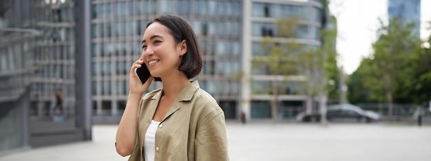 写真 アジアの若いデジタル遊牧民の女の子の垂直ショットは、携帯電話で話し、一緒に通りを歩きます