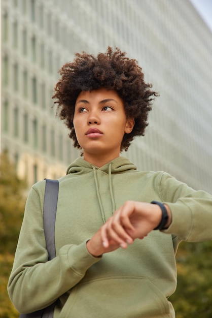 Vertical shot of pensive curly haired young woman in hoodie checks time on wristwatch waits for someone poses with fitness facilities stands against blurred background. Sporty lifestyle concept