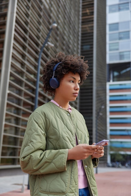 Vertical shot of pensive millennial woman downloads new music tracks holds mobile phone wears stereo headphones on ears enjoys media